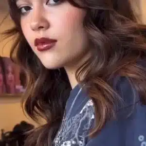 Young woman featuring a layered brunette shag haircut with full bangs and glossy dark red lipstick.