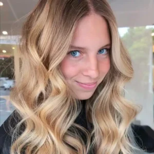 Young woman showcasing long blonde balayage hair with loose waves while wearing a Redken salon cape.