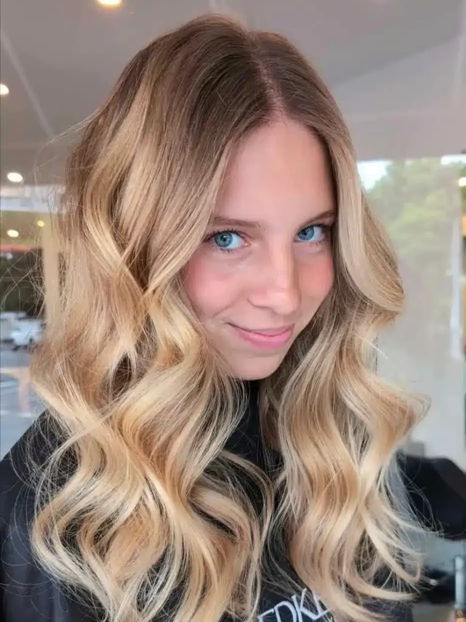 Young woman showcasing long blonde balayage hair with loose waves while wearing a Redken salon cape.
